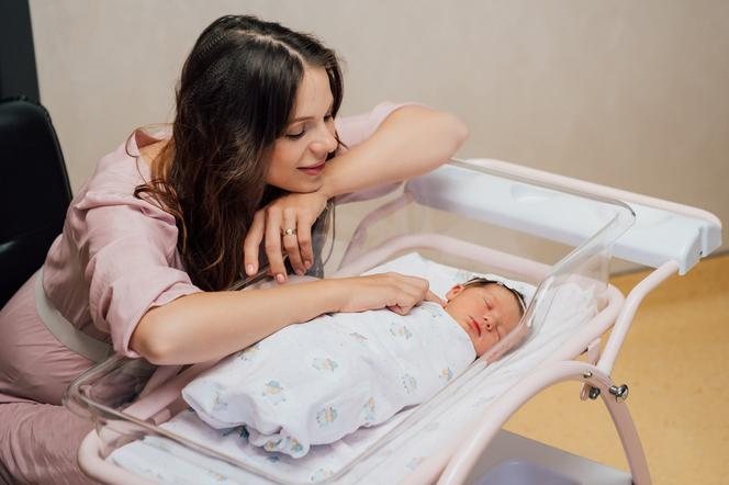 Mama pochyla się nad noworodkiem śpiącym w szpitalnym łóżeczku obok jej łóżka